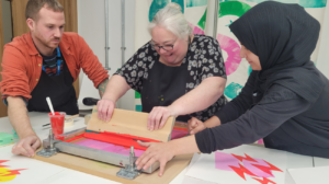 Three people work together on a screen printing project, pressing ink through a stencil onto paper in an art studio.