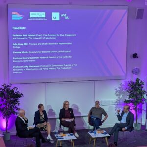 Panel members discuss topic on stage at an event with a powerpoint presentation on the screen behind them
